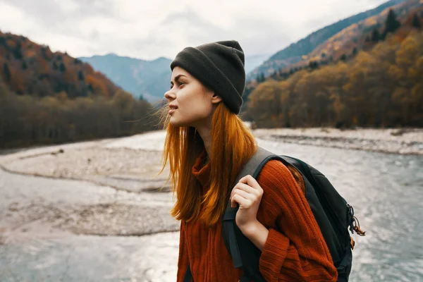 Bela mulher em uma camisola com uma mochila viaja perto de um rio de montanha na vista lateral da natureza — Fotografia de Stock