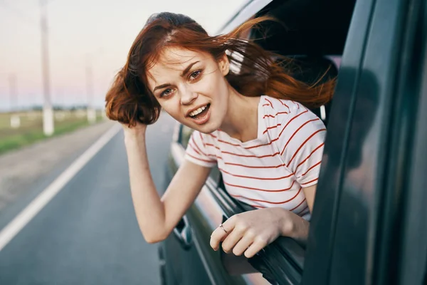 Mulher de cabelos vermelhos em uma camiseta olha para fora da janela do carro e gestos com as mãos na estrada de verão — Fotografia de Stock