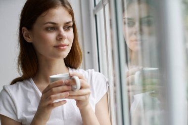 Elinde bardak olan kadın tişörtü. Pencereden içeriye bakıyor.