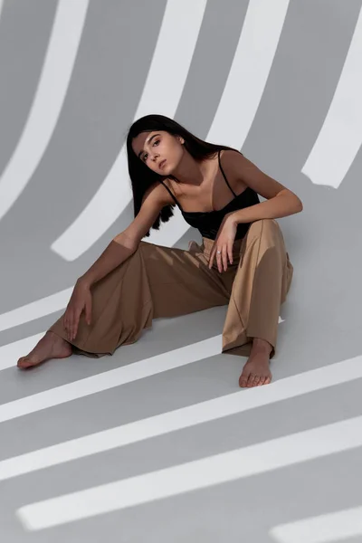 Modna brunetka w beżowych spodniach siedząca na podłodze w pomieszczeniu i padająca cieniem na ścianę — Zdjęcie stockowe