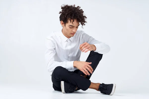 a guy with curly hair in a white shirt, sneakers and trousers sits on the floor In a bright room