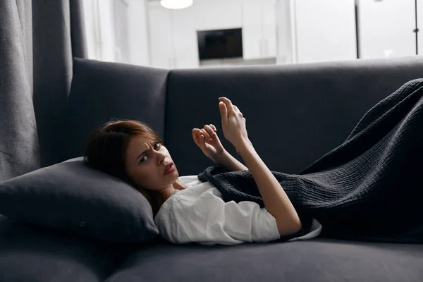 Una mujer se acuesta en un sofá gris cubierto con una manta y un teléfono móvil —  Fotos de Stock
