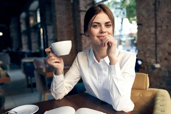 Glad kvinna med en kopp i handen sitter i en stol i restaurangen och interiören i bakgrunden — Stockfoto