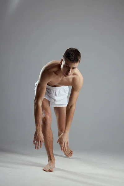 Atleta com um torso bombeado em shorts brancos em um fundo cinza inclinado para a frente dentro de casa — Fotografia de Stock