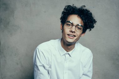 Kıvırcık saçlı gözlüklü ve beyaz gömlekli bir adam kumaş arka planda gülümsüyor yakın plan portresi