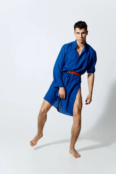 Un hombre bastante gay en un vestido azul con un cinturón está bailando sobre un fondo claro — Foto de Stock