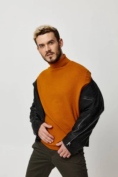 一个金发碧眼的男人，穿着橙色毛衣，穿着皮夹克，手拿裤子，背景浅薄 — 图库照片