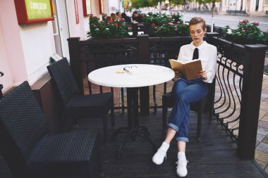 Kafe not defteriyle masada kitap okuyan kadın öğrenci kot pantolonu tişörtü giyiyor.