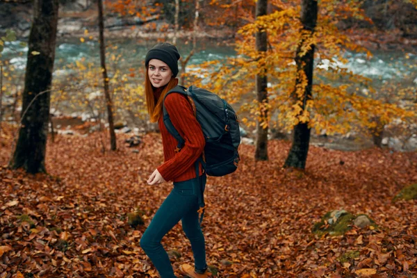 Vrouw in een trui met rugzak en in spijkerbroek wandelt door het herfstbos in de bergen bij de rivier — Stockfoto