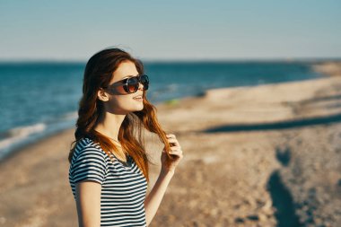 Tişörtlü güneş gözlüklü kadın deniz kıyısındaki kumlu sahilde duruyor.