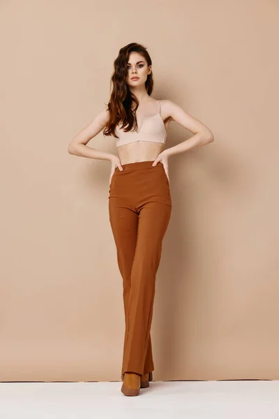 An extravagant woman in trousers and a bra on a beige background holds her hands on her belt — Stock Photo, Image
