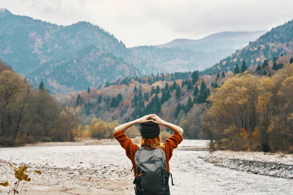 Une voyageuse en pull avec un sac à dos fait des gestes avec ses mains et se détend sur la rive de la rivière dans les montagnes — Photo