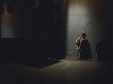 Karanlık bir odada, bir sandalyede oturan yalnız bir kadın. Üzüntü, yalnızlık.