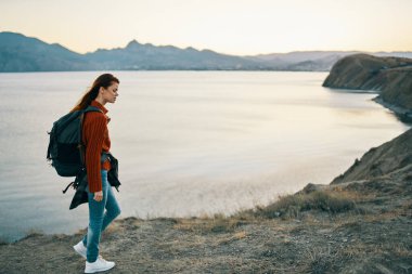 Sırt çantalı, kot pantolonlu ve spor ayakkabılı bir gezgin dağlarda deniz kenarında bir sahilde.