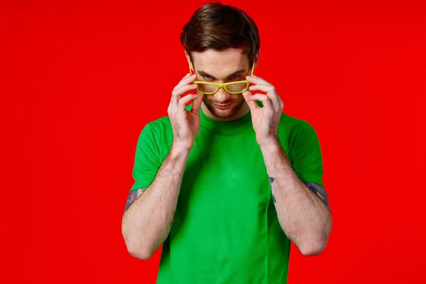 Elinde dövmeleri olan yeşil tişörtlü bir adam kırmızı arka planda gözlük takıyor. — Stok fotoğraf