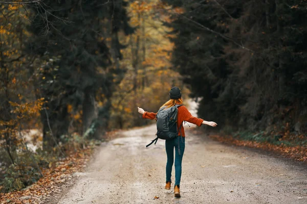 Mulher de casaco jeans com uma mochila caminha ao longo da estrada na floresta de outono — Fotografia de Stock