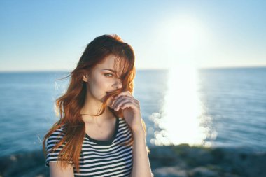 Doğadaki dağlarda gün batımında deniz kenarında tişörtlü kızıl saçlı bir kadın.