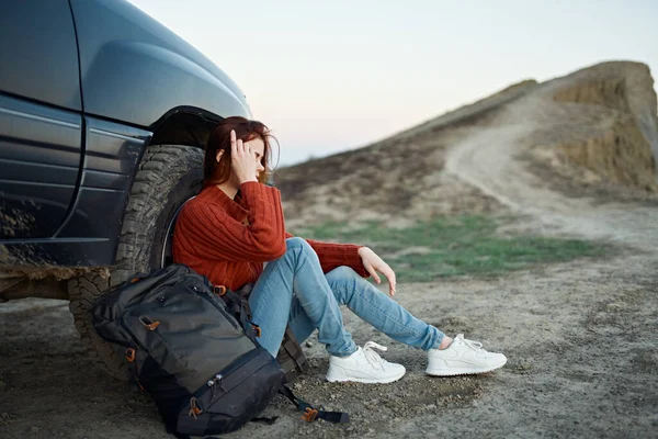 O viajante senta-se perto do carro na natureza nas montanhas e uma mochila no lado — Fotografia de Stock
