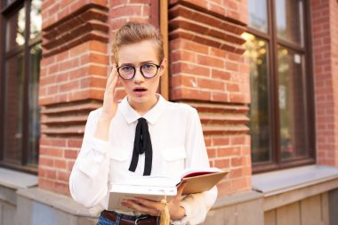 Caddedeki tuğla binanın yanında kısa saçlı gözlüklü bayan öğrenci ve beyaz gömlekli kesim manzaralı.