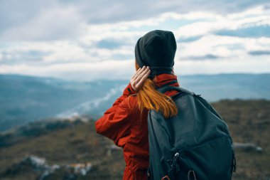 Ceket şapkalı ve sırt çantalı kadın doğadaki dağlara bakıyor.