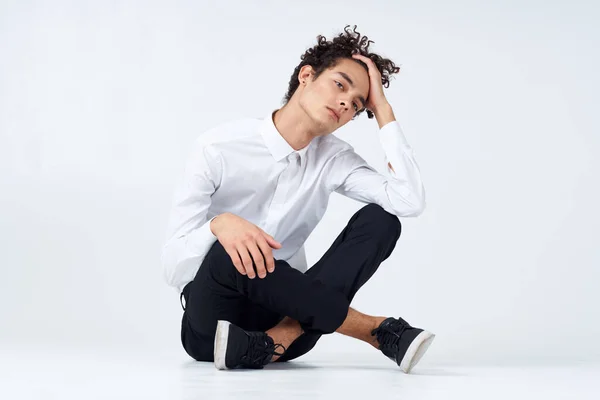 A guy with curly hair in a white shirt, sneakers and trousers sits on the floor In a bright room — Stock Photo, Image