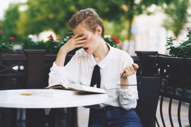 Restoranda bir masada kitap okuyan bir kadın eğitim duyguları arka planda çiçekler gözlüyor.
