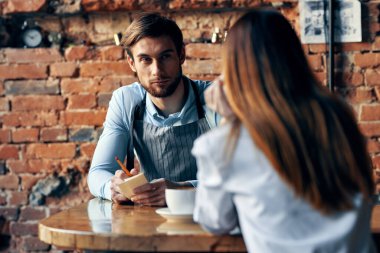Kafe masasında bir fincan kahve ve önlüklü bir adam ve kırmızı etekli bir kadın.