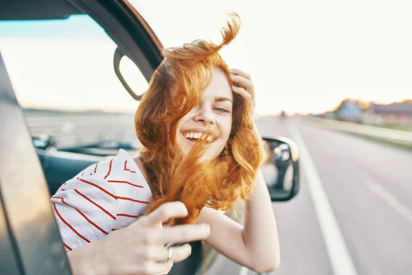 Mujer pelirroja feliz mira por la ventana del coche y conduce en la carretera, pista vacaciones de verano — Foto de Stock