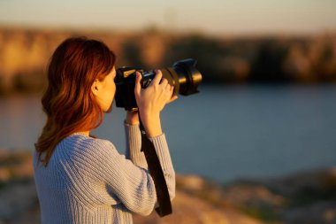 Kadın profesyonel bir kamerayla dağlarda gün batımını deniz kenarında fotoğraflıyor.