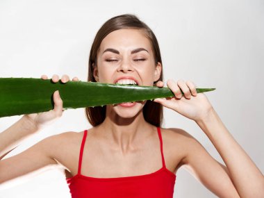 Güzel bir kadın yeşil bir aloe yaprağını kemirdi gözleri kapalı ve açık arkaplanda kırmızı bir tişört giyiyor.