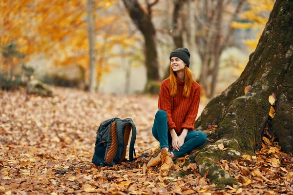 Mulher de calças e botas com uma mochila senta-se perto de uma árvore na floresta de outono folhas caídas — Fotografia de Stock