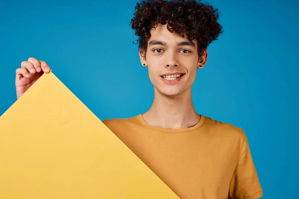 곱슬곱슬 한 노란색 포스터를 갖고 있는 쾌활 한 남자가 파란 배경을 흉내낸다 — 스톡 사진
