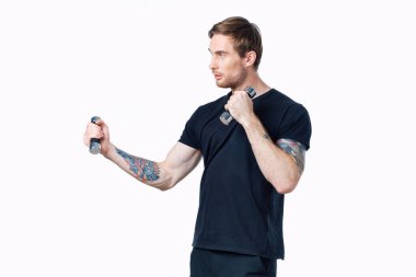 Siyah tişörtlü güçlü bir sporcu ellerinde dambıllarla egzersiz yapıyor.