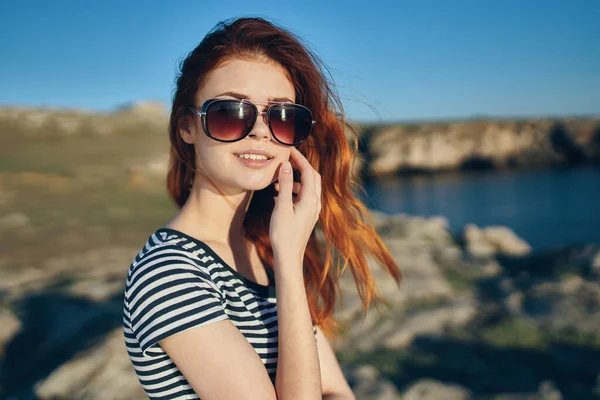 Piękna rudowłosa kobieta w t-shircie i okularach przeciwsłonecznych na łonie natury w górach nad rzeką — Zdjęcie stockowe