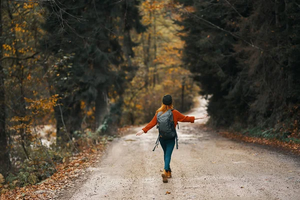 Mujer en chaqueta vaquera con una mochila camina por la carretera en el bosque de otoño — Foto de Stock