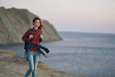 Sırt çantalı bir kadın yürüyüşçü dağlarda deniz kenarında yürüyor.