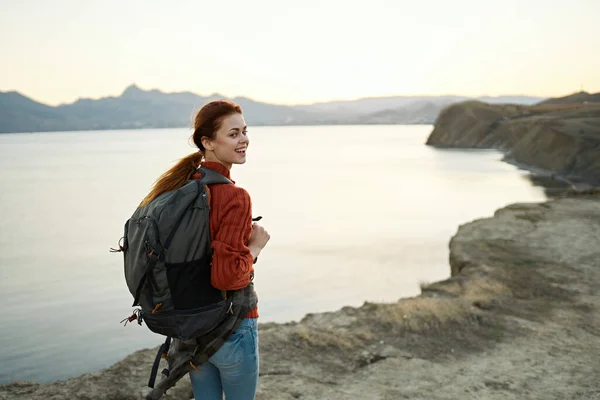 Frau bei Sonnenuntergang am Meer in den Bergen mit einem Rucksack auf den Schultern — Stockfoto