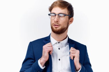 Beyaz gömlekli bir iş adamı ceketini düzeltiyor ve sakallı, açık renk bir arka planı var.