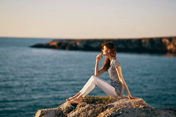 A woman in a striped t-shirt and white trousers sits on the beach near the sea in the mountains — Stock Photo, Image