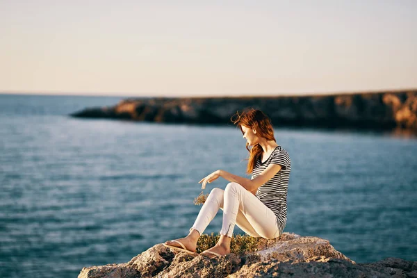 Beyaz pantolonlu ve tişörtlü bir kadın dağlarda deniz kenarında bir taşın üzerinde oturuyor. — Stok fotoğraf