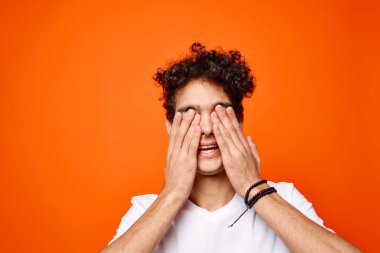 Kıvırcık saçlı, beyaz tişörtlü adam duygularını turuncu arka planda tutuyor.