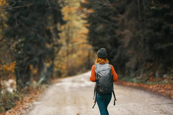 Mulher com uma mochila em um chapéu e uma camisola laranja na estrada na floresta de outono — Fotografia de Stock