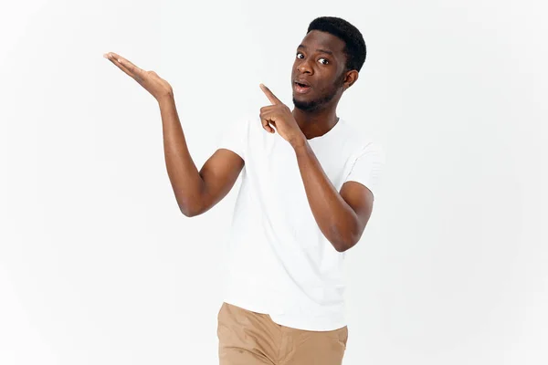 Un chico guapo de apariencia africana gestos con las manos sobre un fondo claro y señala su dedo hacia un lado — Foto de Stock