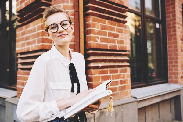 Žena v blízkosti cihlové budovy v brýlích s knihou v rukou na ulici vzdělávání — Stock fotografie