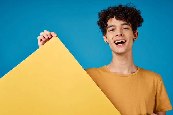 손에 노란 포스터를 들고 파란 배경을 광고하는 곱슬머리의 감정적 인 남자 — 스톡 사진