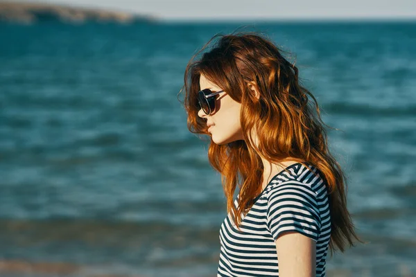 Donna dai capelli rossi in occhiali da sole sulla spiaggia vicino al mare blu e le montagne sullo sfondo — Foto Stock