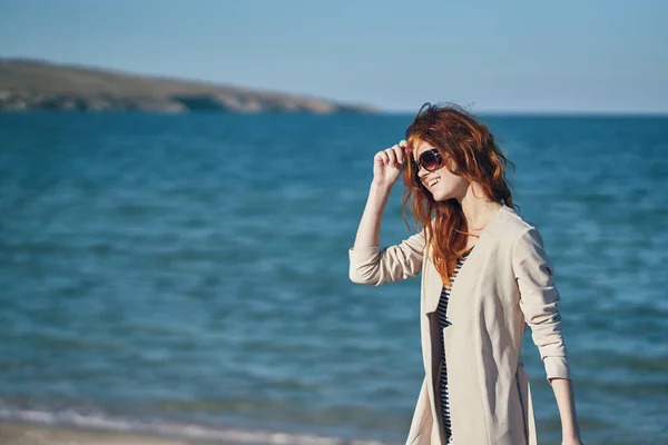 Red-haired traveler in a coat and a t-shirt on the beach near the sea summer vacation — Stock Photo, Image