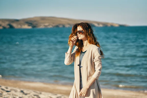 Mujer en la arena en la playa cerca del mar en un abrigo turismo gafas de sol —  Fotos de Stock