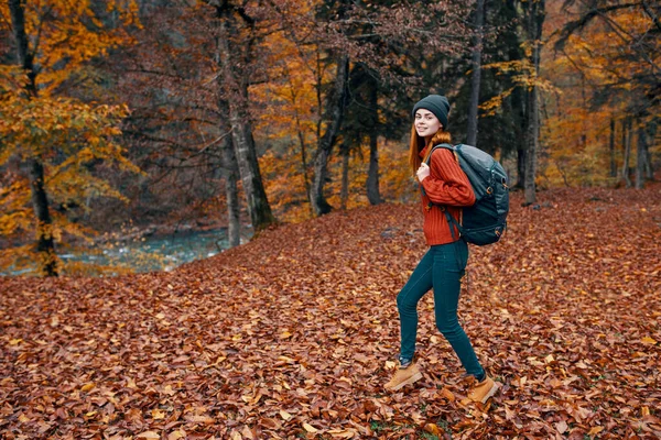快乐的女旅行家背着背包在大自然的秋日公园里散步，在河边的风景旁边，高大的树毛衫 — 图库照片