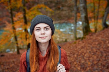 Kırmızı kazak ve şapkalı sırt çantalı enerjik bir turist parktaki sonbahar ormanında dinleniyor.